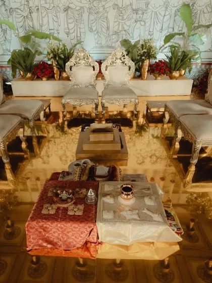 The seating arrangement for the couple and close family inside the mandap. The ornate silver and gold chairs are placed on a reflective golden floor, surrounded by traditional ritual items and floral decor.