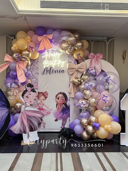 A magical ballerina and butterfly theme for a first birthday party. This elaborate setup features large purple butterfly wing props, ballerina character standees, a light-up marquee number '1', and a balloon arch in pastel purple and gold.