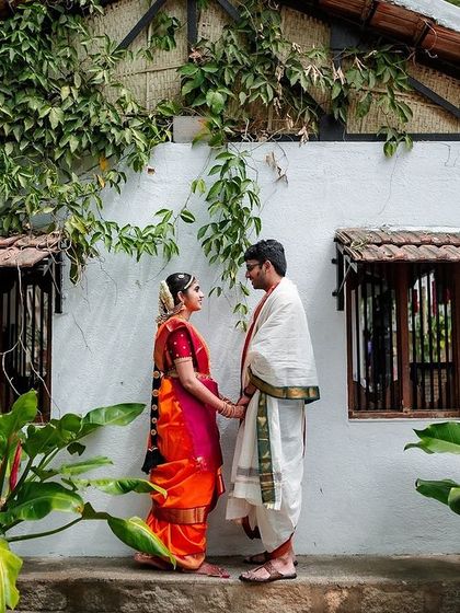A quiet portrait of a couple in traditional South Indian wedding attire, holding hands in front of a rustic, charming house.