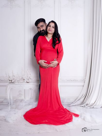 A full-length portrait of a couple against a classic white paneled wall. The mom-to-be's red gown provides a beautiful pop of color.