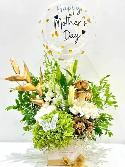 An elegant Mother's Day arrangement in a gift box. It features a mix of white and green flowers, golden accents, and a "Happy Mother's Day" balloon.