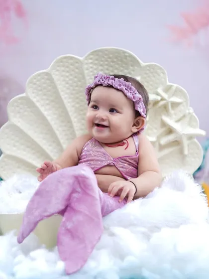 A happy baby girl in a lavender mermaid costume, smiling brightly from her seashell throne.
