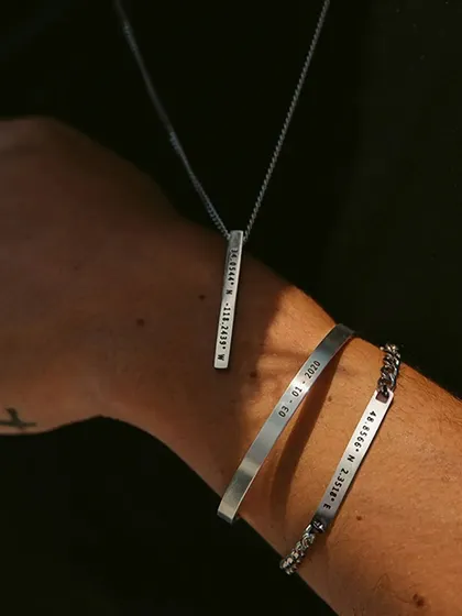 A collection of personalized jewelry, including a cuff bracelet with a date, a chain bracelet with coordinates, and a bar necklace. This shows how you can stack different meaningful pieces.