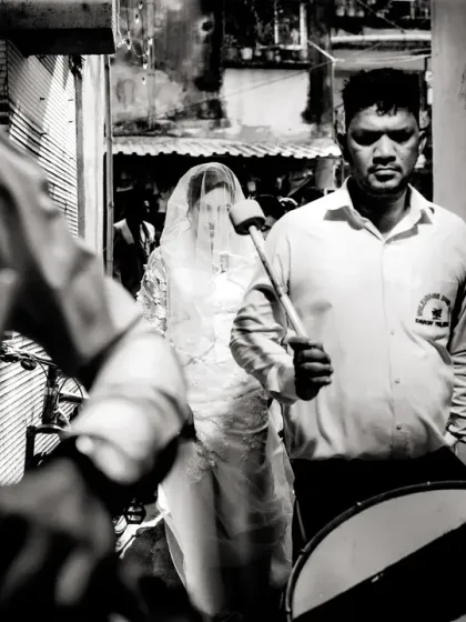 A black and white street photography style shot, capturing the bride walking behind the wedding band.