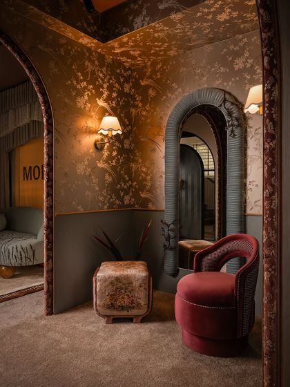 This composition highlights the interplay of different shapes and textures. The arched mirror, framed in pleated fabric, contrasts with the curved velvet chair and the floral ottoman, all set against a backdrop of delicate wallpaper.