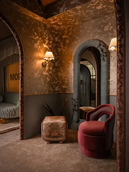 This composition highlights the interplay of different shapes and textures. The arched mirror, framed in pleated fabric, contrasts with the curved velvet chair and the floral ottoman, all set against a backdrop of delicate wallpaper.