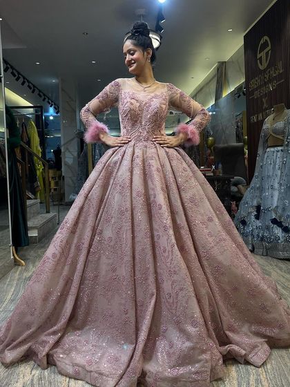 A full-length view of a client smiling in the dusty pink, long-sleeved ball gown. The feather cuffs add a touch of playful glamour to this elegant and sparkling princess dress.