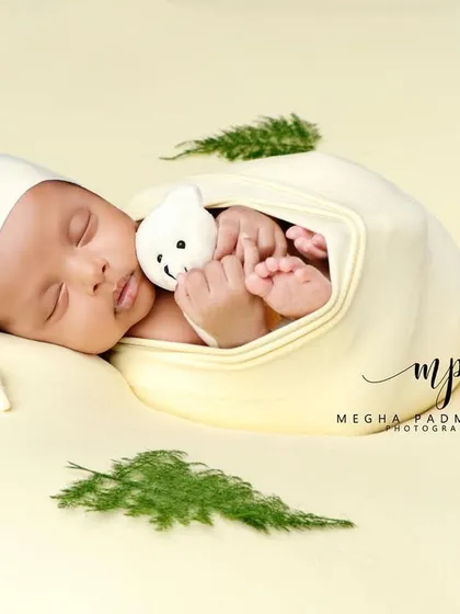 A baby in a pale yellow wrap and cap, cuddling a tiny toy. The green leaves add a touch of nature.