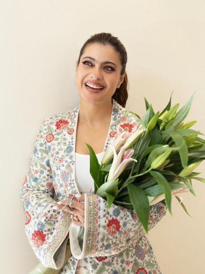 Kajol looking radiant in a floral embroidered jacket and pants set, holding a bouquet of fresh lilies.