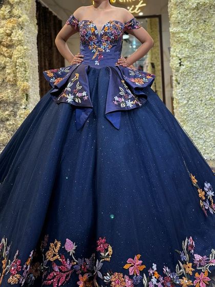 A client trying on a navy blue ball gown with colorful floral embroidery. This front view shows the flattering off-shoulder design and unique peplum waist.