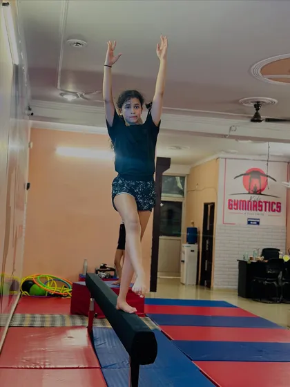 A student demonstrates perfect form and balance on the beam. Her arms are raised high as she prepares for her next move, showing great concentration.