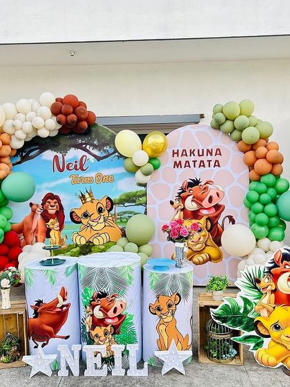 A complete Lion King 'Hakuna Matata' themed birthday setup. It features multiple character cutouts, themed plinths, and an organic balloon garland in jungle-inspired colors.