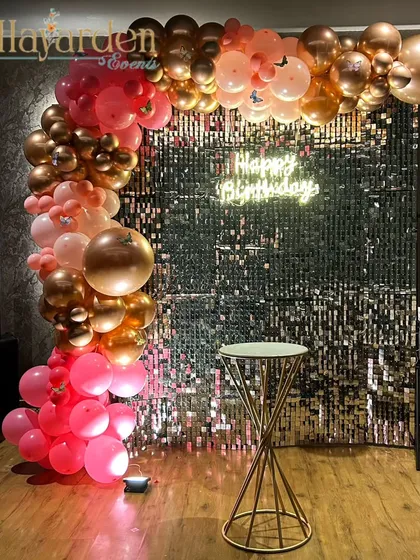 A chic and modern party setup with a rose gold and pink balloon arch against a sequin backdrop. The 'Happy Birthday' neon sign and minimalist pedestal create a stylish look.