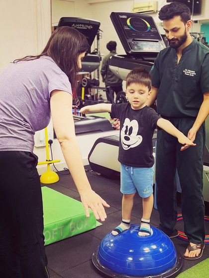 A therapist and parent encourage a young boy as he practices balancing on a BOSU ball. This collaborative approach ensures the child feels supported from all sides.