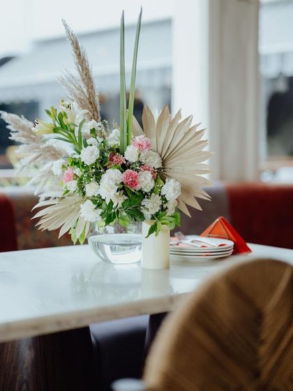 A close-up of a table centerpiece, featuring pampas grass, dried palm leaves, and soft pink and white flowers in a simple glass vase.