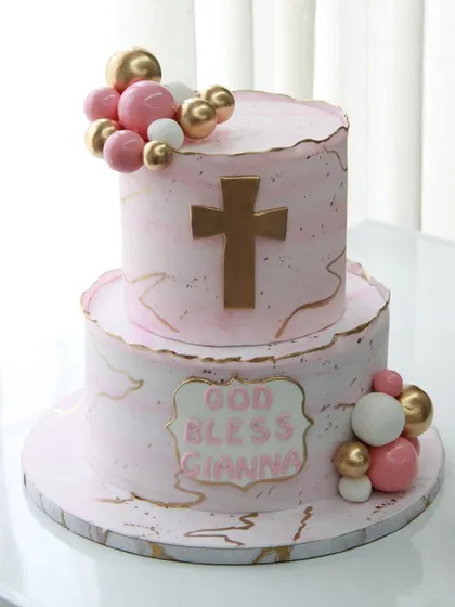 "God Bless Gianna." A beautiful two-tiered baptism cake with a pink marble effect, gold accents, and a cross.