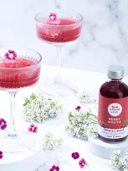 An elegant, high-key shot of Bab Louie & Co.'s Berry Mojito mixer with prepared cocktails. The styling with fresh flowers creates a delicate and sophisticated look for the beverage.