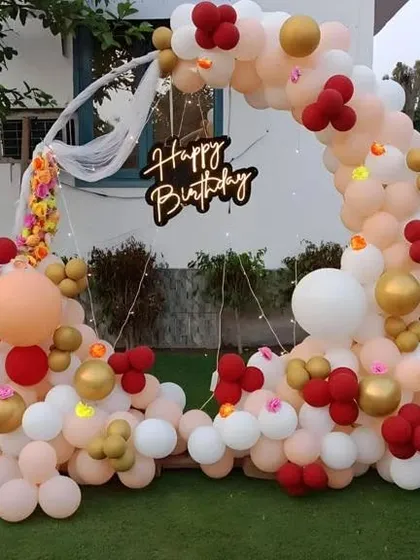 An outdoor birthday party with a circular balloon arch in red, white, and peach, and a 'Happy Birthday' neon sign.