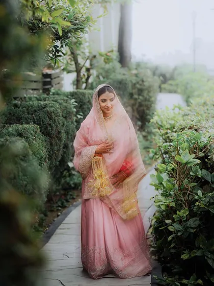 A bride in a soft pink suit walks through a green pathway, a moment of quiet grace before the ceremony.