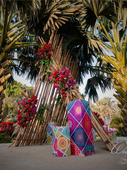 A unique seating area for a Mehendi, featuring a triangular bamboo structure and chairs with vibrant, geometric patterns, set against the backdrop of palm trees.