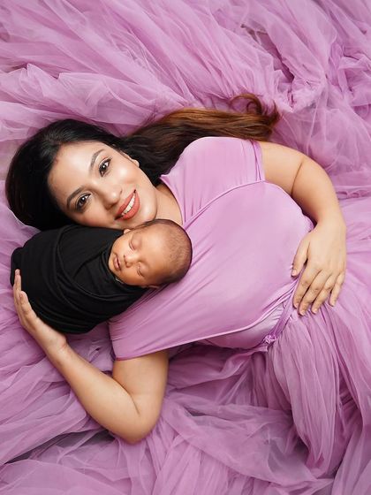 A mother's serene moment with her newborn. Lying on a bed of soft purple fabric, she cradles her sleeping baby, creating a dreamy and artistic portrait.