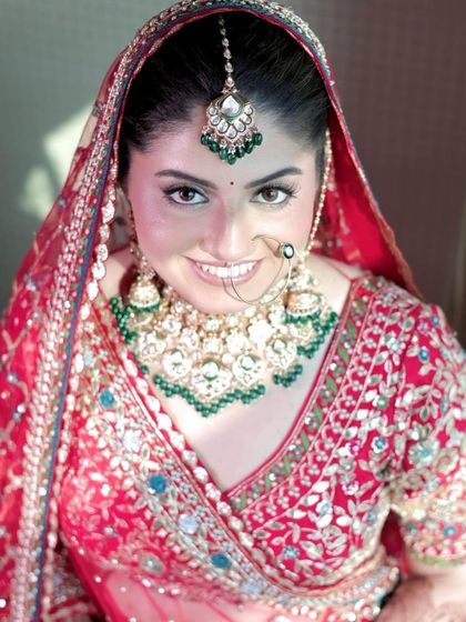 A close-up of a smiling bride, her face framed by a red dupatta and a stunning green-beaded jewellery set.
