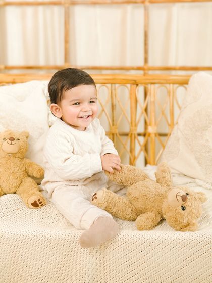 A classic sitter session portrait. This baby's bright, happy smile is captured beautifully in a simple, light-filled studio setting with a rattan bench and some teddy bear friends.