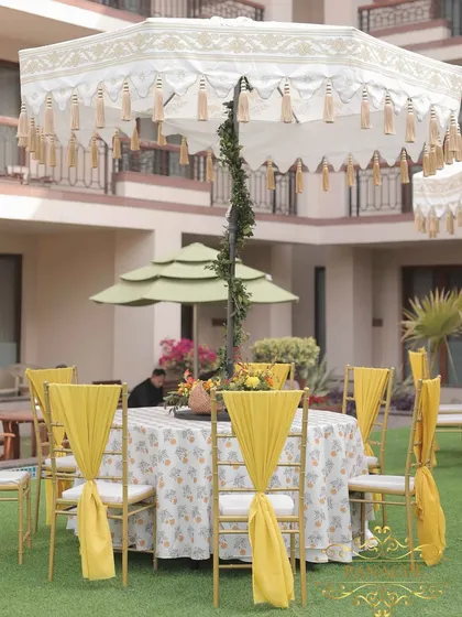 Guest seating for the Haldi, with printed tablecloths, yellow chair drapes, and large decorative umbrellas providing shade and style.