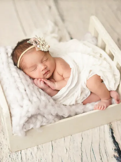 A newborn girl, adorned with a delicate headband, sleeps peacefully in a miniature white bed. I use soft, chunky knit blankets to add texture and warmth to my newborn portraits.