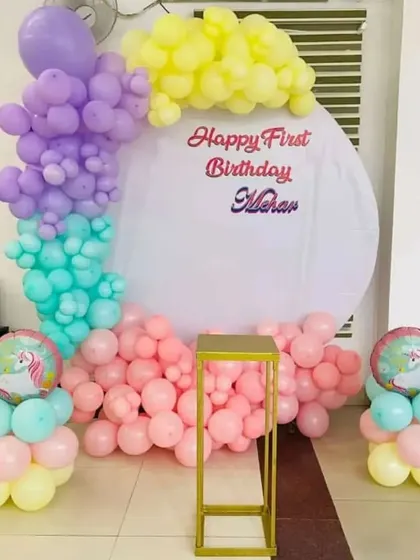 A pastel rainbow theme for a first birthday. This decor includes a circular backdrop with a custom message and a balloon arch in soft shades of purple, yellow, teal, and pink, with unicorn balloon accents.