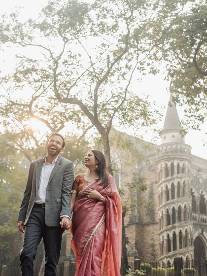 A candid moment of laughter while walking through a park, with the beautiful gothic architecture of a Mumbai university in the background.