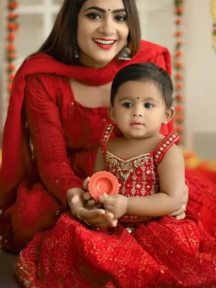 A beautiful mother and daughter portrait from a Diwali mini session. Both are dressed in festive red, with the baby holding a small diya, set against a backdrop of bright, celebratory decorations.