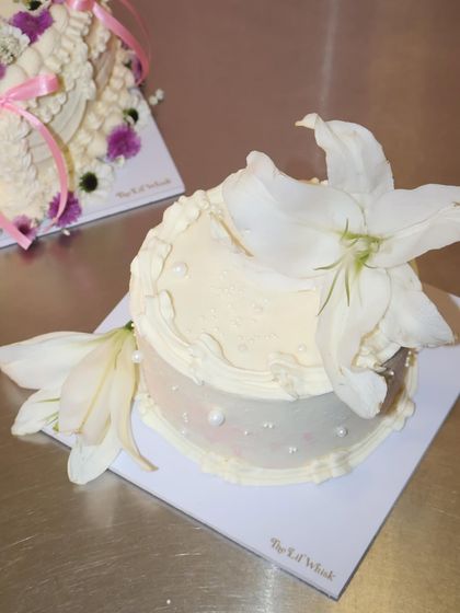 For a soft girl, minimalist vibe, this simple white cake is decorated with elegant piping, pearl details, and two beautiful fresh white lilies.