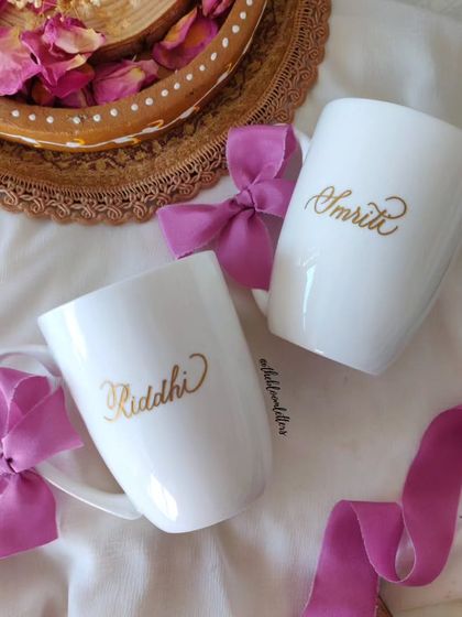 A perfect gift for friends or sisters. These white ceramic mugs are personalized with the names 'Riddhi' and 'Smriti' in a beautiful gold script, complete with delicate pink ribbon accents.