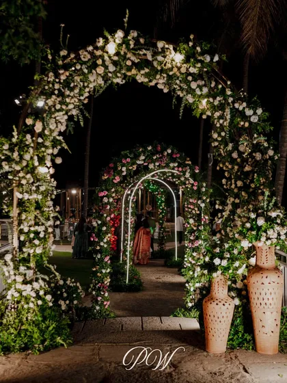 A magical, multi-arched floral entrance for a night event. The layers of arches adorned with white flowers and lights create a sense of depth and wonder as guests walk through.