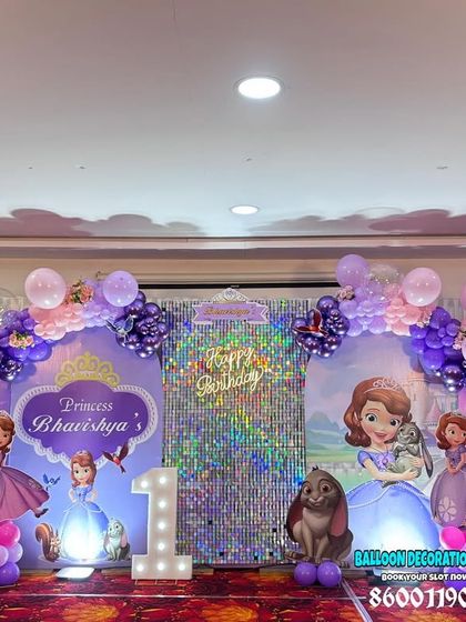 A complete Princess Sofia theme for a first birthday party. We created a wide stage setup with multiple character cutouts, a shimmer backdrop, and purple and pink balloon arches.