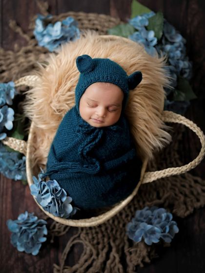 A little bear cub sound asleep. This newborn is wrapped snugly in a basket, surrounded by blue flowers, creating a dreamy and adorable portrait.