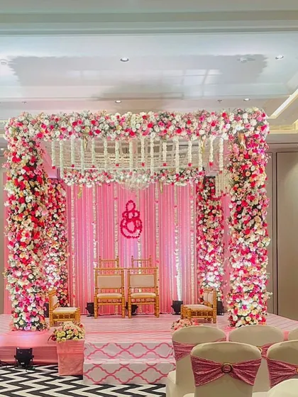 An elegant indoor mandap design featuring pillars heavily decorated with pink and white flowers. The monogram of the couple adds a personalized touch to the sacred space.