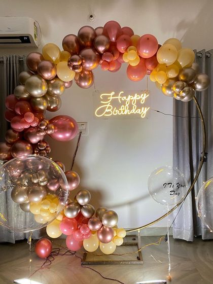 This is a duplicate of ID 17, 49, 153, 186, 209. An elegant balloon ring decoration for an intimate home celebration. This setup features a half-arch of chrome and pastel balloons on a golden ring, with a 'Happy Birthday' neon sign.