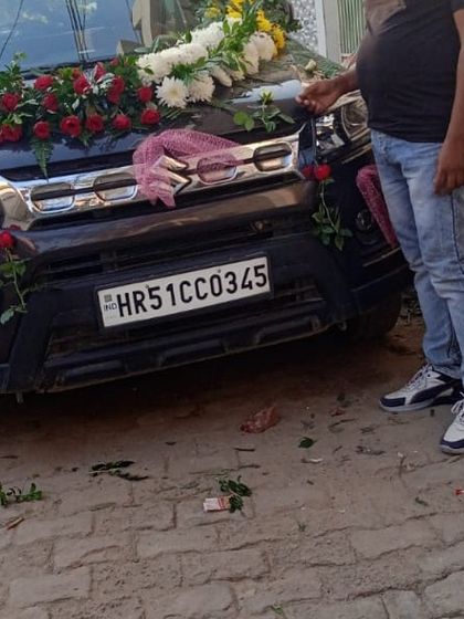 A black Maruti Suzuki Brezza with a floral garland and pink ribbons. This image shows the decoration in progress.