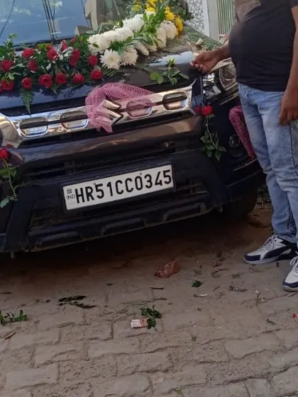 A black Maruti Suzuki Brezza with a floral garland and pink ribbons. This image shows the decoration in progress.