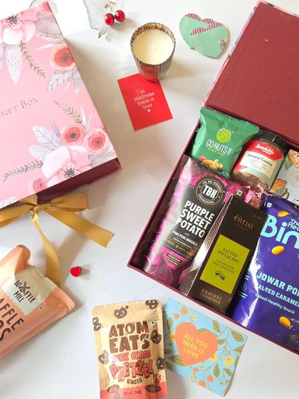 A top-down view of a Valentine's Day hamper overflowing with goodies. It includes a mix of sweet and savory snacks like waffle chips and purple sweet potato crisps, all packed in a lovely floral box.