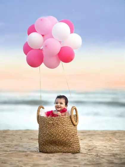 A wider shot of the magical hot air balloon setup on the beach, showing the baby happily floating above the sand against a sunset sky.