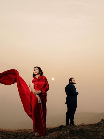 An artistic pose during a sunset couple shoot. The wind catches the bride's red saree, creating a sense of movement and elegance against the soft light of the setting sun.