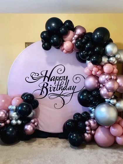 This unique birthday backdrop features a custom printed pink circular panel with 'Happy Birthday' in elegant calligraphy. An organic arrangement of black, pink, and silver chrome balloons completes the modern look.