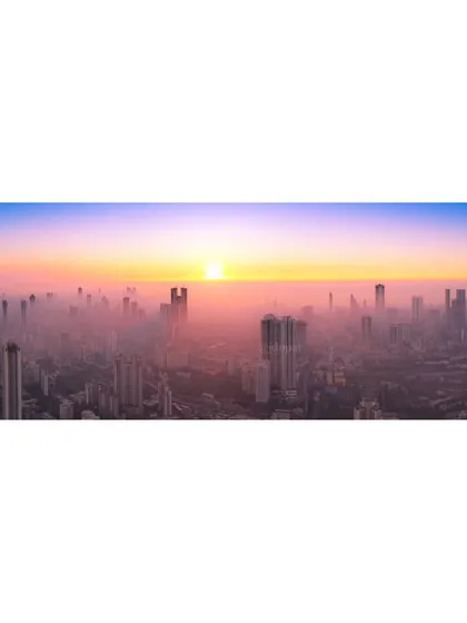 A panoramic drone photograph of Mumbai's hazy skyline at sunset, capturing the vast expanse of the city under a colorful sky.