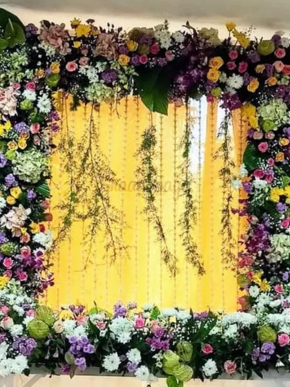 This vibrant floral frame is bursting with a variety of colorful flowers, including purple, yellow, and white chrysanthemums and roses. The hanging green elements in the center add a touch of rustic charm to the setup.