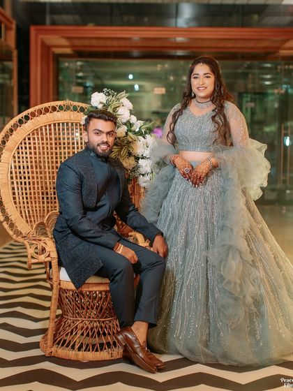 A regal portrait of the couple at their engagement ceremony. The groom is seated on a peacock chair while the bride stands beside him, both looking elegant and happy.