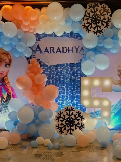 A magical 'Frozen' themed birthday party for a 5 year old. The backdrop features Elsa and Anna, surrounded by a balloon arrangement in shades of blue, white, and peach, with snowflake accents.