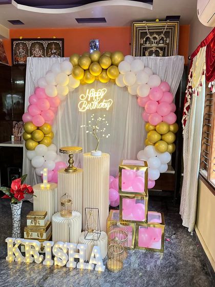 A lovely home birthday decoration in pink, white, and gold. This setup includes a balloon arch, a white curtain backdrop, a 'Happy Birthday' neon sign, and various props like bird cages and gift boxes.